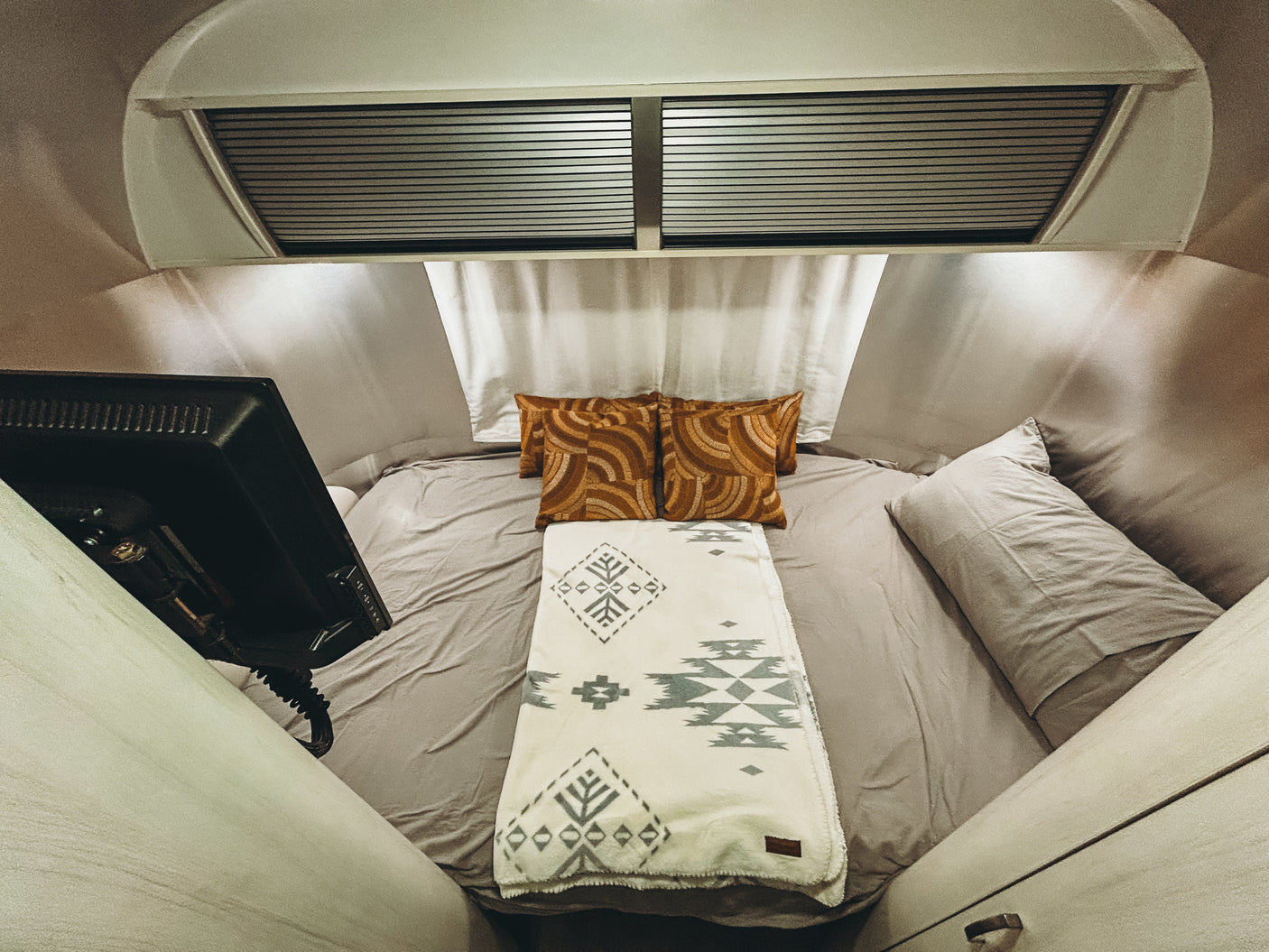 Inside a comfy RV rental bedroom: grey bed, orange patterned pillows, geometric blanket, and TV. Ideal for road trip nights.