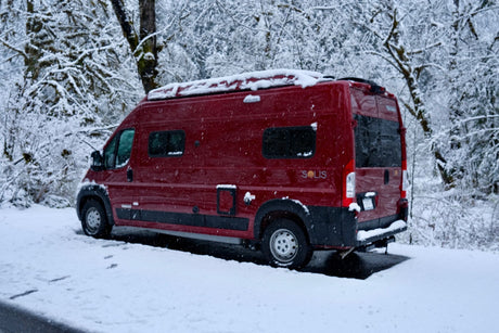 A vibrant red Winnebago Solis camper van is nestled in a snowy winter forest, perfect for an RV rental adventure.
