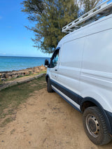 Enjoying coastal views from a white RV rental van parked on sandy ground by the calm blue ocean.