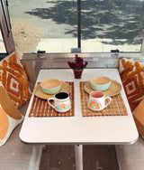 Cozy RV rental dining space with two settings, wood plates, mugs on bamboo mats, and a scenic window view.