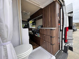 RV rental interior: compact bathroom with toilet/shower, dark wood cabinets, and living area featuring a car seat.