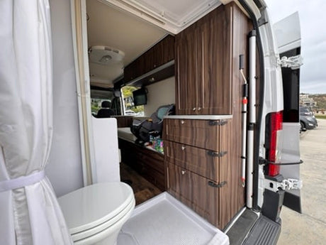 RV rental interior: compact bathroom with toilet/shower, dark wood cabinets, and living area featuring a car seat.