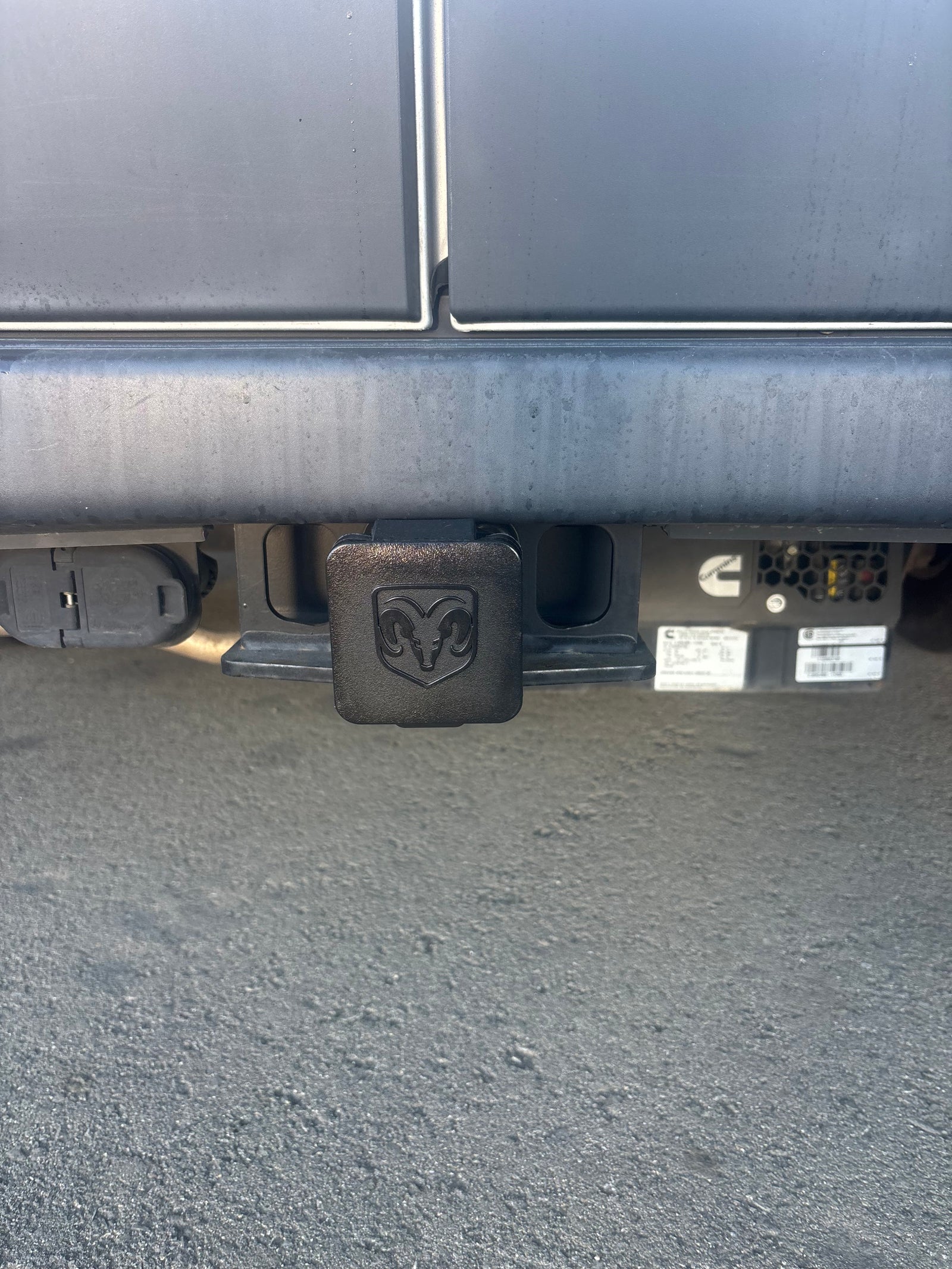Rear view of an RV rental's trailer hitch with a Ram logo cover, trailer wiring connector, and a Cummins generator.