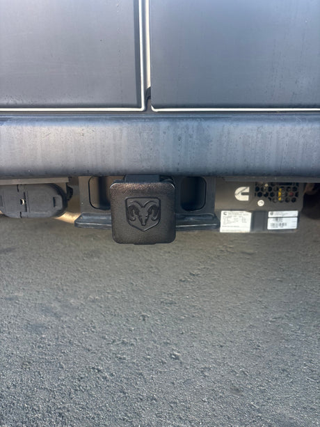 Rear view of an RV rental's trailer hitch with a Ram logo cover, trailer wiring connector, and a Cummins generator.