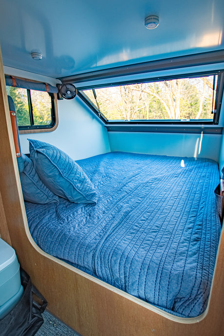 An inviting blue bed inside a compact RV rental, featuring windows with natural views and a ceiling fan.