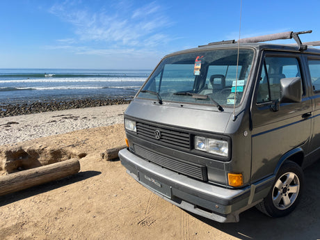 A gray VW camper van, ideal for RV rental, parked on a sunny beach by the ocean with waves and surfers in the distance.