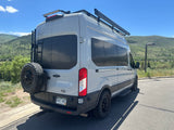 This rv rental, a light grey Ford Transit camper van with a roof rack and awning, is parked against a scenic mountain backdrop.