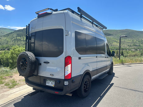 This rv rental, a light grey Ford Transit camper van with a roof rack and awning, is parked against a scenic mountain backdrop.