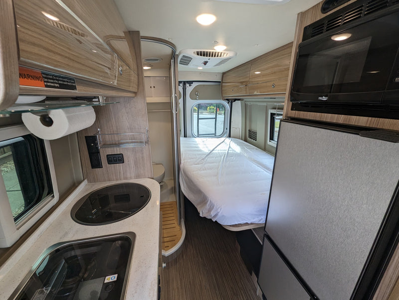  parked on a street beside a vibrant autumn park.|This silver Winnebago Travato camper van (RV rental) is parked on a sunny road with autumn trees and hills in the background.|This RV rental's interior features a compact kitchen with sink/stovetop — photo 3