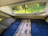 Cozy RV rental: bed with blue comforter & patterned blanket, large window view of vibrant green nature.