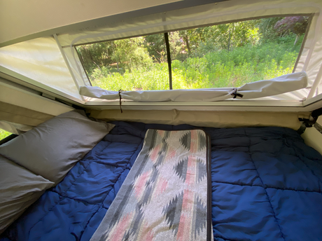 Cozy RV rental: bed with blue comforter & patterned blanket, large window view of vibrant green nature.