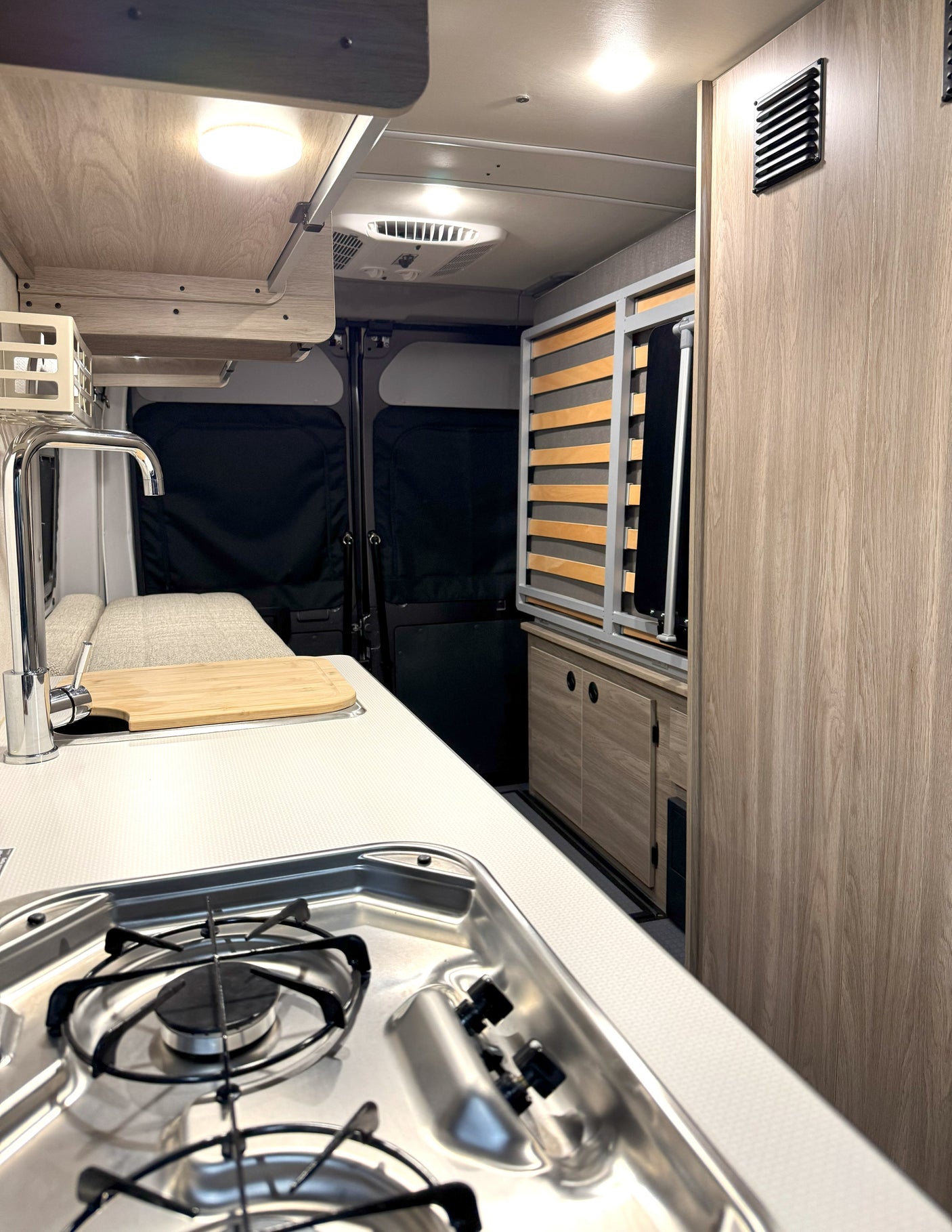 Inside a modern rv rental: kitchenette with stove, sink, cutting board, wooden cabinets, and a slatted bed base.