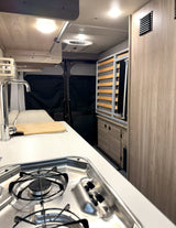 Inside a modern rv rental: kitchenette with stove, sink, cutting board, wooden cabinets, and a slatted bed base.