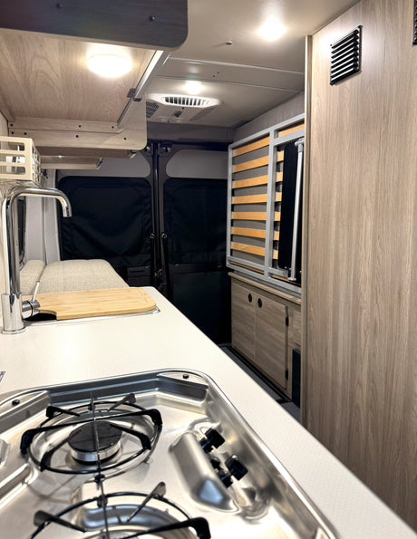 Inside a modern rv rental: kitchenette with stove, sink, cutting board, wooden cabinets, and a slatted bed base.