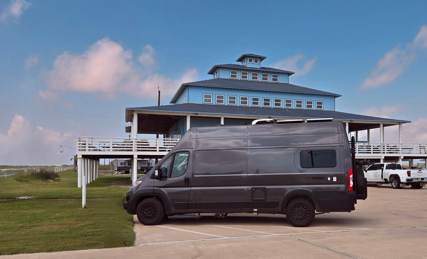 Dark gray Noovo rv rental van parked at a large blue building. Explore travel & vacation possibilities.