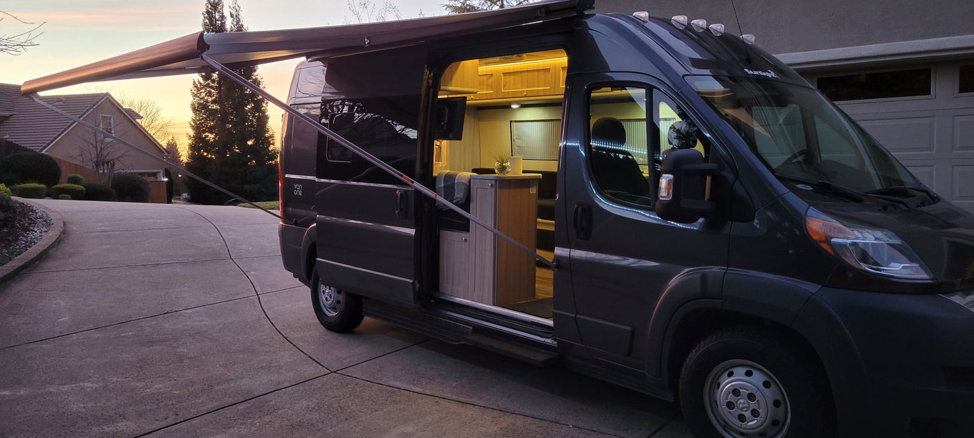 Dark gray rv rental camper van with an extended awning. Its brightly lit, inviting interior is visible through the open side door at sunset.