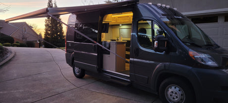 Dark gray rv rental camper van with an extended awning. Its brightly lit, inviting interior is visible through the open side door at sunset.