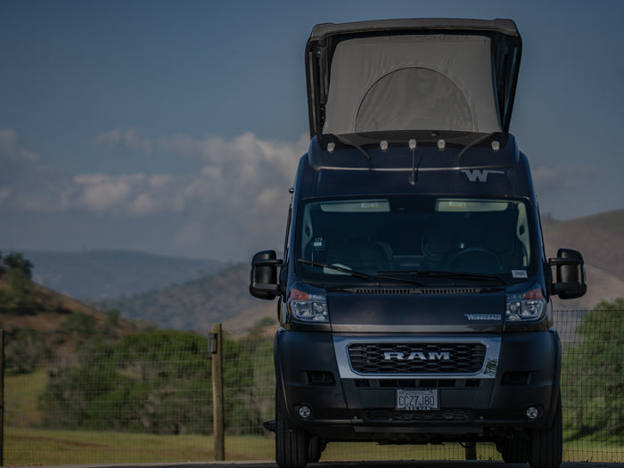 A dark Winnebago Ram ProMaster rv rental van, with its pop-top extended, is parked against a scenic backdrop of hills.