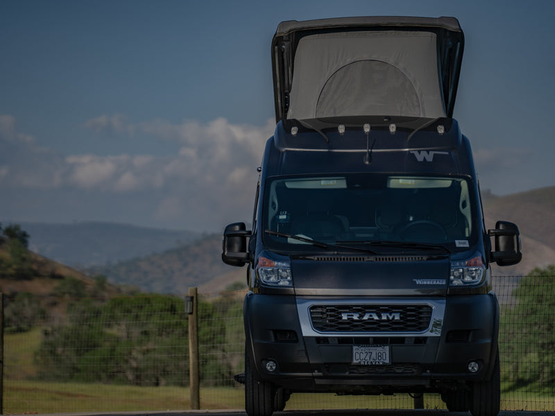 A dark Winnebago Ram ProMaster rv rental van, with its pop-top extended, is parked against a scenic backdrop of hills. — photo 5