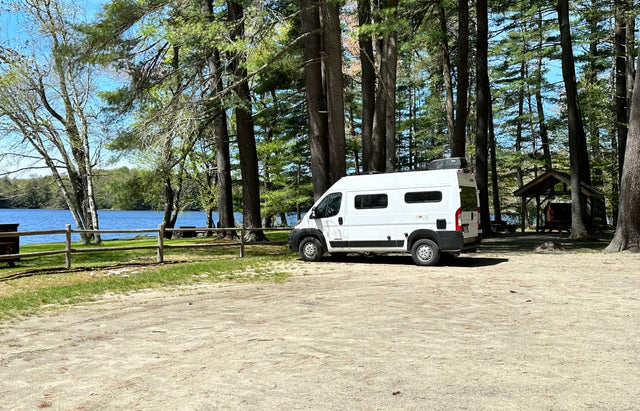 A white RV rental camper van is parked at a scenic lakeside campsite among tall pines