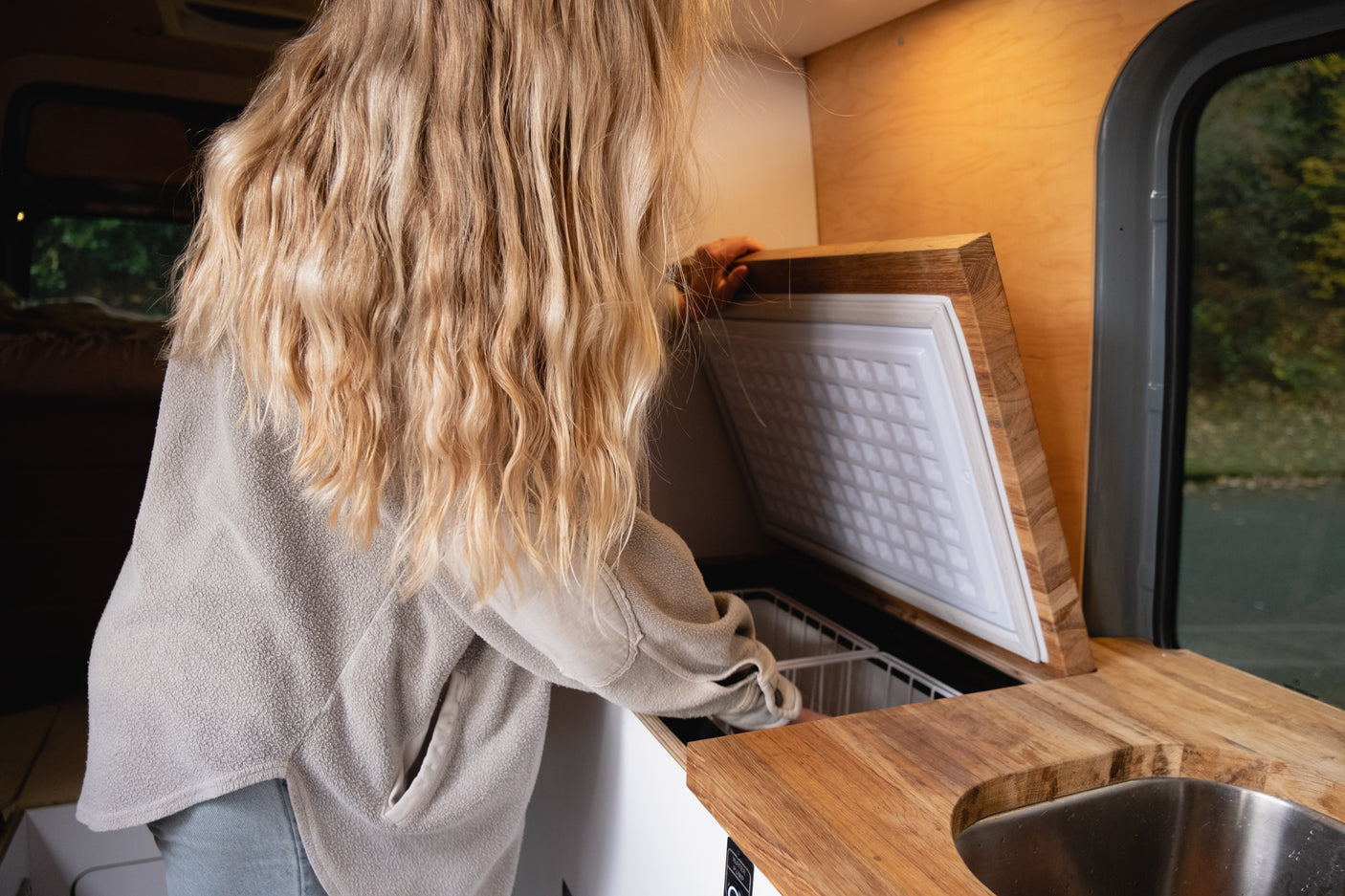 A person with blonde hair reaches into a built-in top-load fridge in an rv rental van's wooden kitchen.