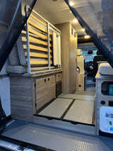 Explore this modern RV rental interior, showcasing sleek cabinetry, a convertible bed, and clever storage in this camper van conversion.