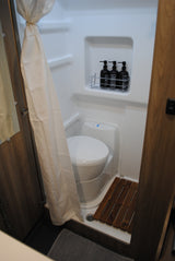 RV rental's compact bathroom: shower, toilet, wooden floor mat, and labeled bath product bottles.
