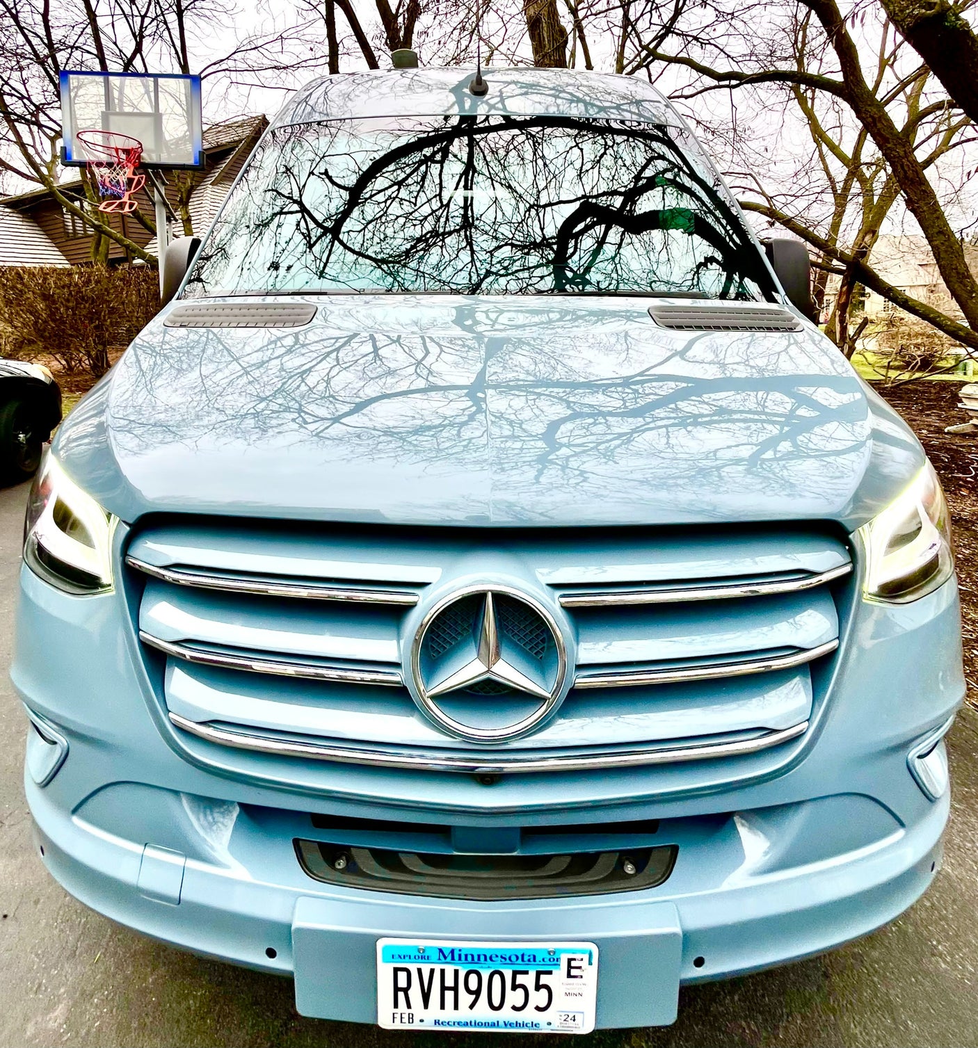 Front view of a light blue Mercedes-Benz Sprinter camper van, ideal for RV rental. Bare trees reflect on its sleek surface.