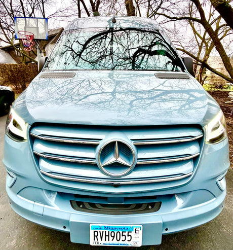 Front view of a light blue Mercedes-Benz Sprinter camper van, ideal for RV rental. Bare trees reflect on its sleek surface.