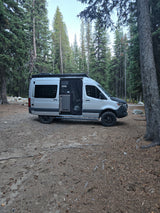 Silver camper van RV rental parked on a dirt site surrounded by tall evergreen trees. Ideal for forest camping.