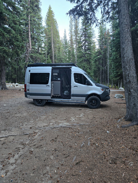 Silver camper van RV rental parked on a dirt site surrounded by tall evergreen trees. Ideal for forest camping.