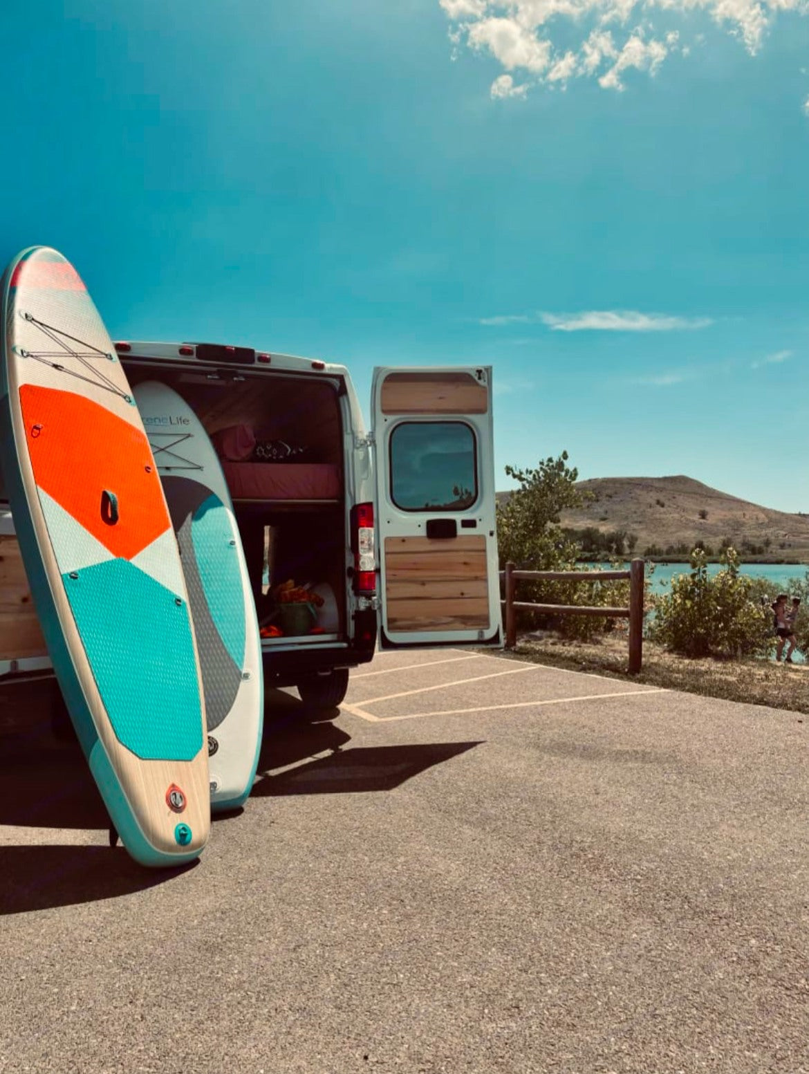 An RV rental, a white camper van, stands with colorful paddleboards by a blue lake, perfect for outdoor adventure.