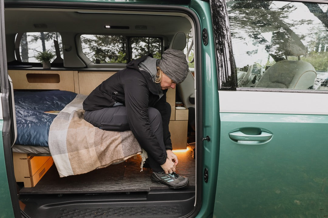 Inside a green rv rental camper van, a person in outdoor gear sits on the bed tying their shoe, preparing for adventure.