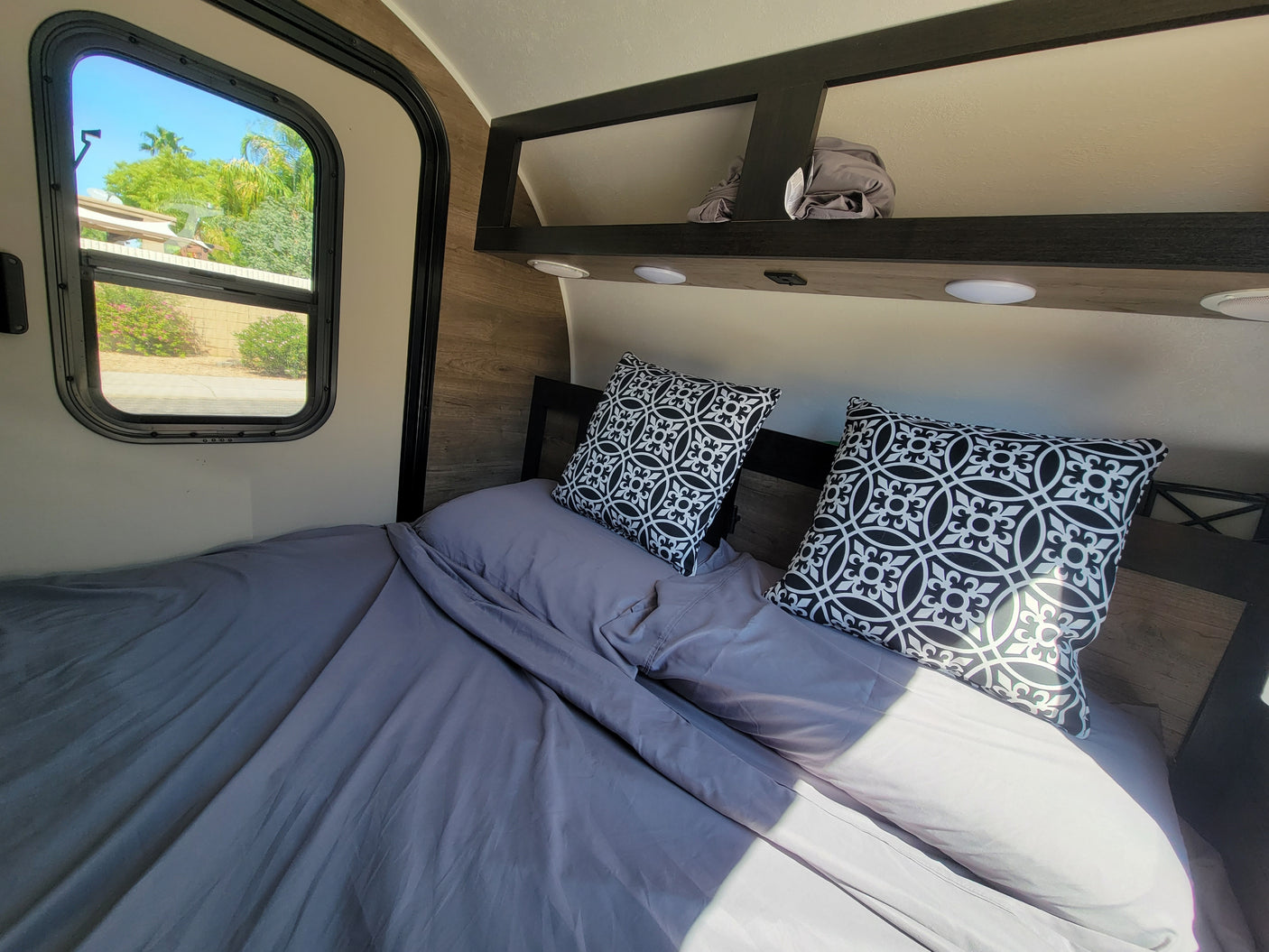 Inside an RV rental: a bed with gray linens, decorative pillows, window to sunny outdoors, and overhead storage.