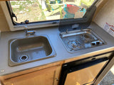 This RV rental kitchenette features a compact sink, 2-burner stove, and under-counter refrigerator, ideal for travel.