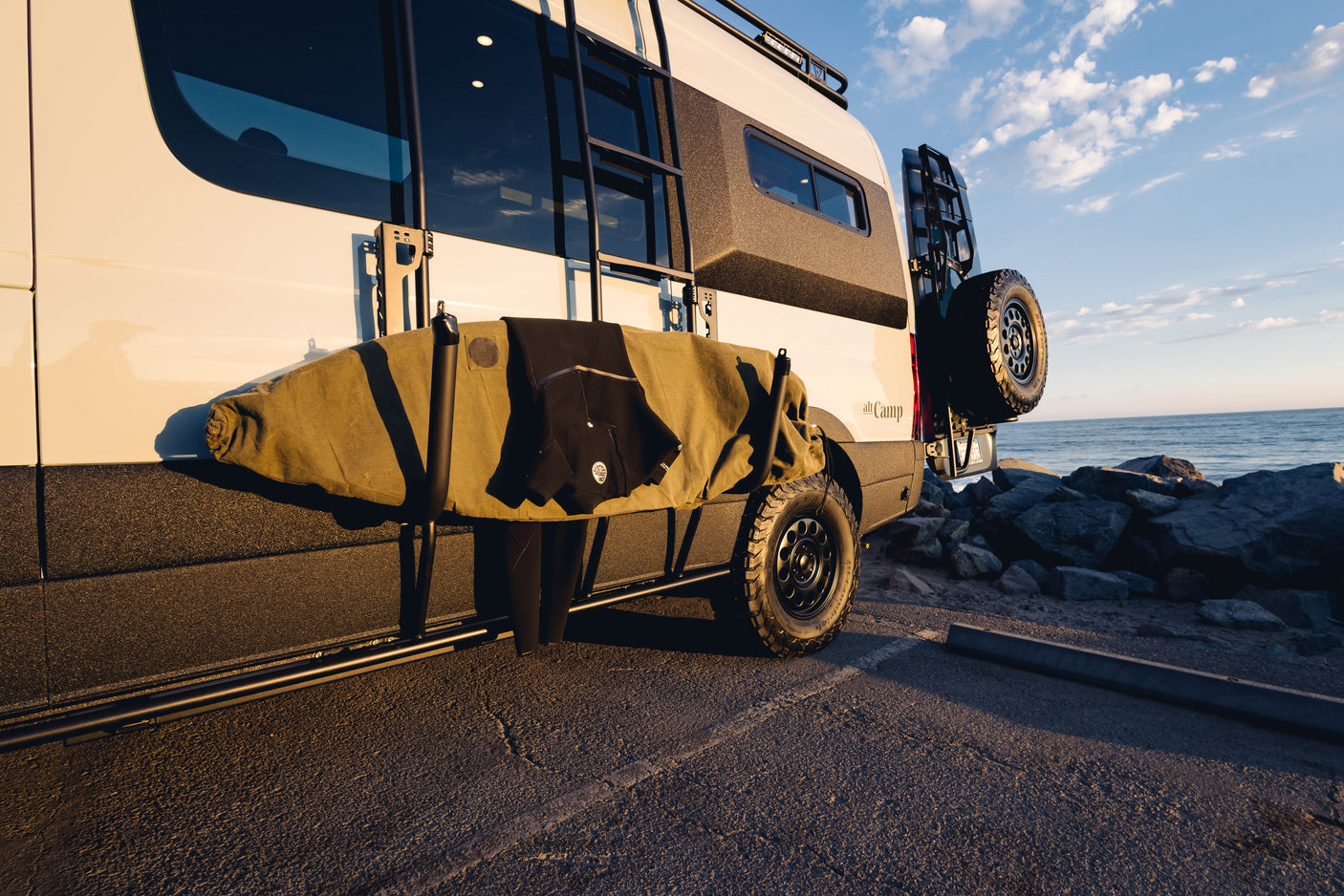 This RV rental camper van with a surfboard and wetsuit is parked by the ocean at sunset, ready for adventure.