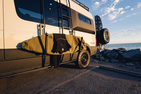 This RV rental camper van with a surfboard and wetsuit is parked by the ocean at sunset, ready for adventure.