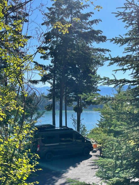 Discover nature's beauty: a black RV rental parked by a sparkling lake, framed by green trees and mountains.