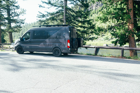 A dark gray Flatlander camper van, an ideal rv rental, by a scenic river and towering trees. Perfect for adventure travel.