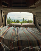 Cozy RV rental bed with a striped quilt, offering a scenic view of evergreen trees, islands, and ocean through the open back doors.