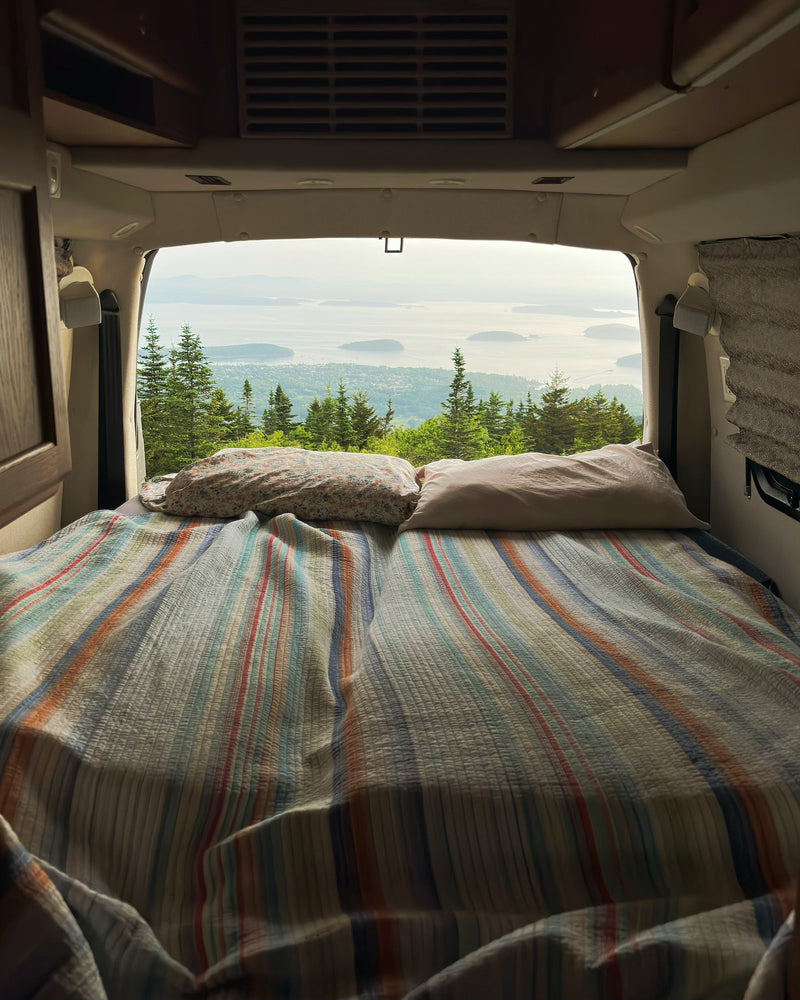 Cozy RV rental bed with a striped quilt, offering a scenic view of evergreen trees, islands, and ocean through the open back doors. — photo 2
