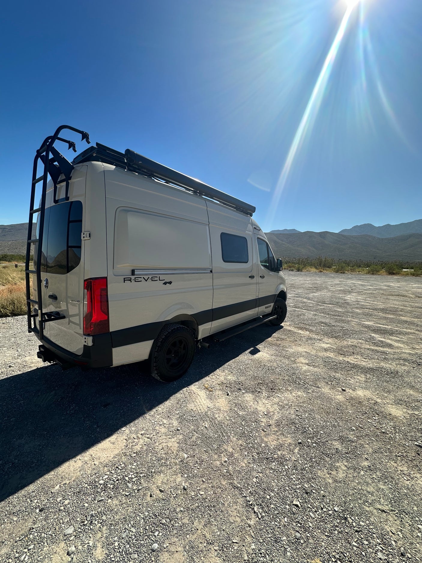 RV rental: Winnebago Revel 4x4 camper van parked on gravel, ready for adventure under a bright, sunny sky with mountains.