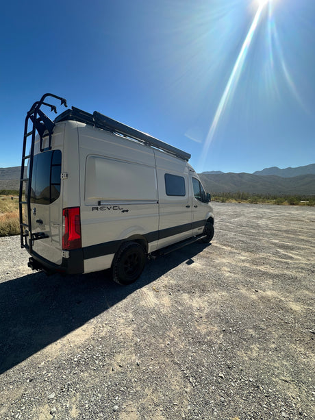 RV rental: Winnebago Revel 4x4 camper van parked on gravel, ready for adventure under a bright, sunny sky with mountains.