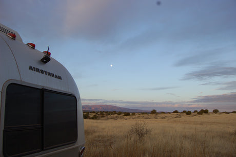 An Airstream RV rental is seen from behind, parked in a vast desert landscape at dusk under a faint moon.