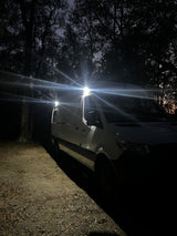 An rv rental camper van with bright roof lights illuminates a dark, wooded campsite at night, showcasing its readiness for adventure.