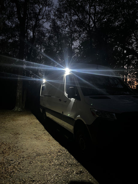 An rv rental camper van with bright roof lights illuminates a dark, wooded campsite at night, showcasing its readiness for adventure.