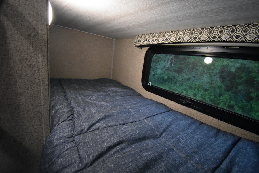 RV rental sleeping area: cozy bed with blue comforter, light, and window revealing lush green trees. — photo 14