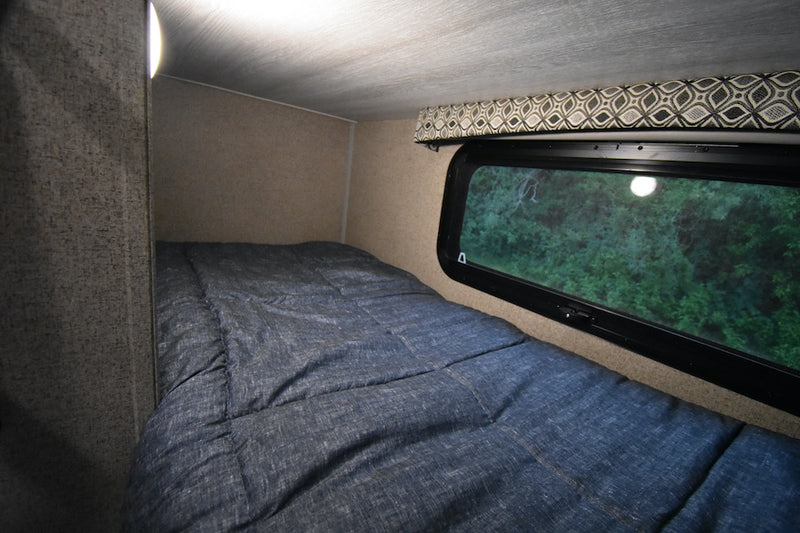 RV rental sleeping area: cozy bed with blue comforter, light, and window revealing lush green trees. — photo 14
