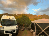 An RV rental camper van and a large screen tent are set up in a mountain campsite at sunset with clouds above.