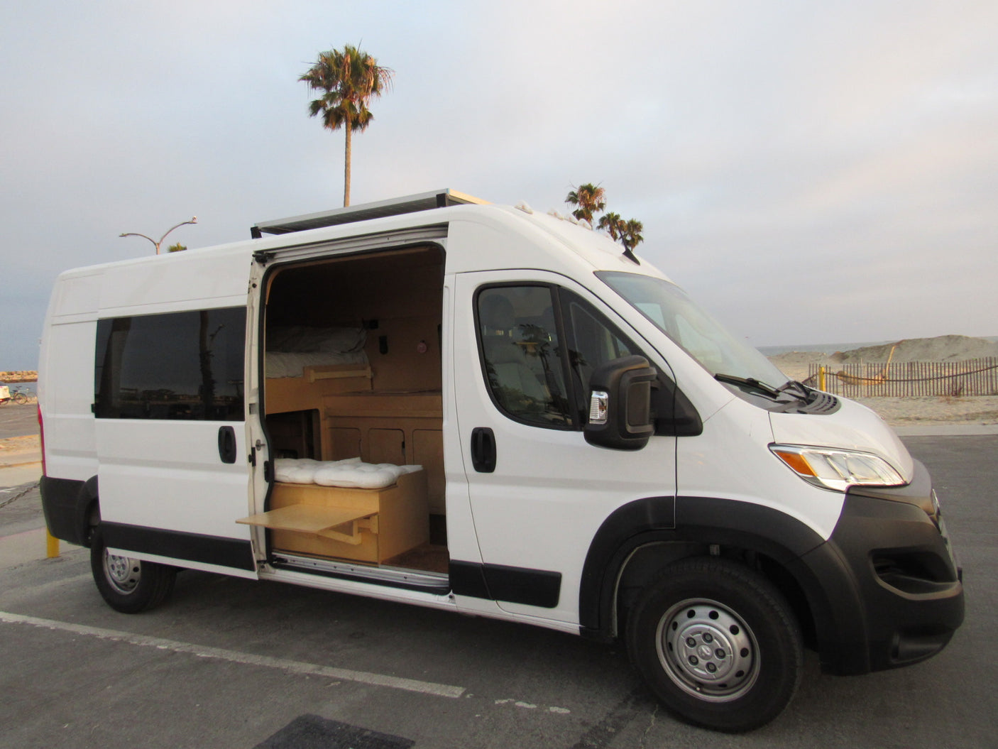 White RV rental camper van with an open side door, showing a custom wood interior, parked near a sandy beach with palm trees.